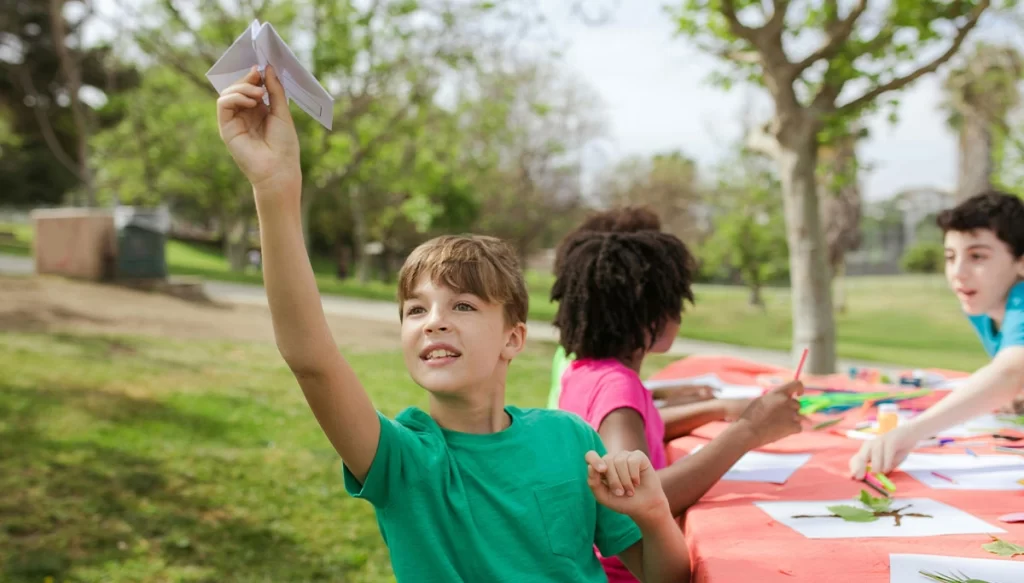 young boy flying a paper plane while sitting at a desk with other students outdoors